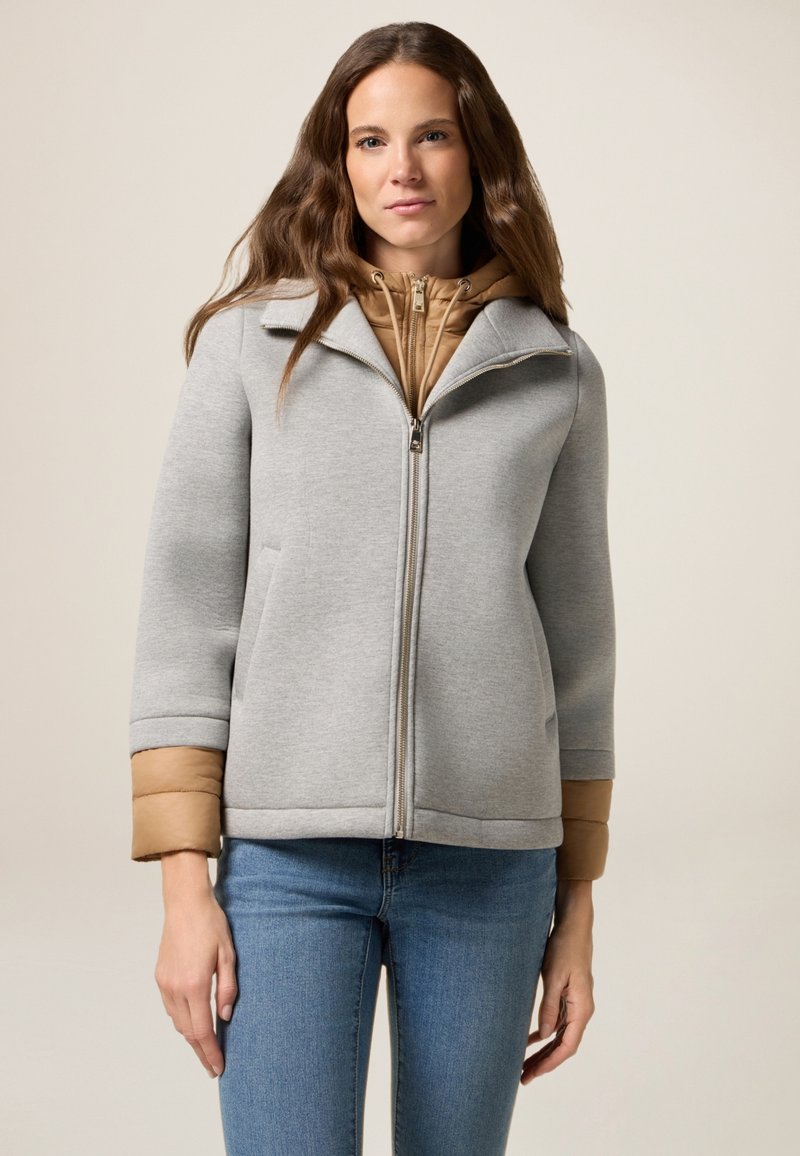 Giacca grigia con zip, colletto e dettagli beige sui polsini, caratterizzata da una texture liscia e da un design strutturato. Indossata sopra uno strato con cappuccio marrone.