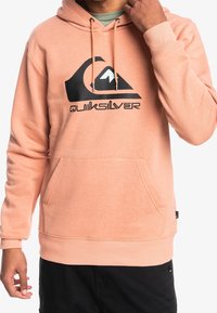 Persikkavärinen huppari, jossa musta logopainatus. Valmistettu pehmeästä kankaasta, siinä on etutasku, naruhuppu ja ribatut hihansuut.