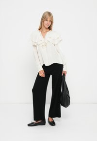 Witte blouse met asymmetrische ruches, knoopsluiting aan de voorkant; gecombineerd met zwarte wijde broek en zwarte flats; met een zwarte tas.
