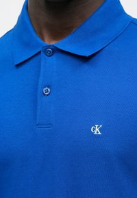 Kék pamut póló texturált szövettel, kéttollú gombolós megoldással és kis fehér "cK" logóval a bal mellrészen.