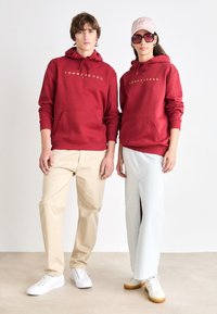 Sudaderas rojas con el logo "TOMMY JEANS", combinadas con pantalones beige y una falda de mezclilla clara. Los accesorios incluyen una gorra rosa y gafas de sol de gran tamaño.