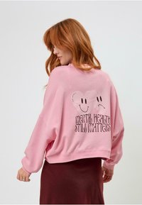 Rózsaszín, oversized kapucnis pulóver mosolygós arccal és a hátán a "MENTAL HEALTH STILL MATTERS" felirattal; puha anyag, laza szabás, lejtős vállakkal.