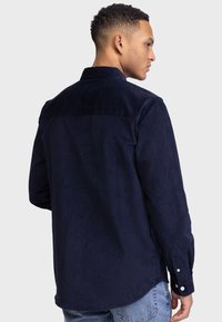 Camisa de pana en color azul marino con cuello, mangas largas y puños con botones. Textura suave con una silueta estilizada.