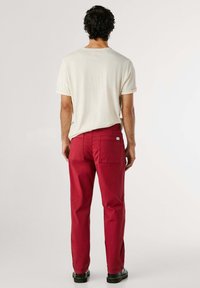 Červené bavlnené nohavice s vreckami na zadnej strane, v kombinácii s krémovým tričkom s krátkym rukávom. Celý outfit dopĺňajú čierne topánky.