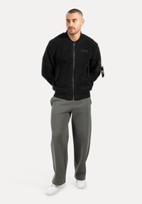 Chaqueta bomber de forro polar negra con cierre de cremallera, puños y dobladillo acanalados. El modelo lleva pantalones de chándal grises y zapatillas blancas. Presente un detalle de marca menor.