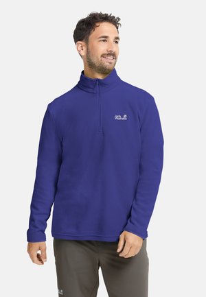 Uomo che indossa un maglione in pile blu Jack Wolfskin con mezza zip e pantaloni scuri, che guarda leggermente verso destra con un leggero sorriso.