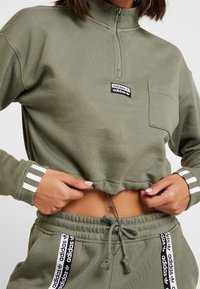 Olivgrön kort tröja med halvlång dragkedja vid kragen, med framficka, varumärkespåse och vita randiga muddar. Mjuk tygstruktur.