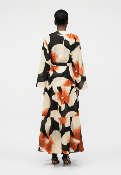 Vrouw met donker haar in een lage knot draagt een lange, vloeiende zwarte jurk met grote witte en oranje bloemenprints, gecombineerd met zwarte pumps.