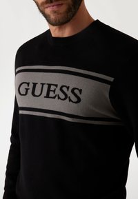 Fekete pulóver, vízszintes szürke sávval, amelyen a "GUESS" szó vastag betűkkel szerepel; bordázott nyakkivágással és texturált anyaggal.