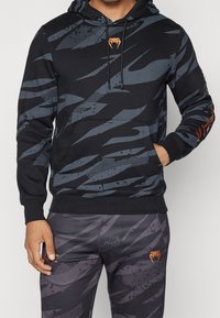 Zwarte hooded sweatshirt met grijze abstracte strepen, voorzien van een kangaroo-zak aan de voorkant en een oranje logo op de borst en de mouw.