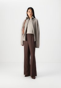 Pull en tricot beige côtelé sous un cardigan gris ample, associé à un pantalon large marron et des chaussures marron, complété par un sac à épaule marron.