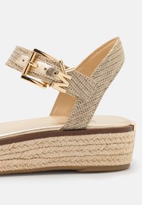 Gros plan d’une sandale compensée beige tissée avec une bride texturée, une boucle dorée et un détail de semelle en corde tressée sur fond blanc.