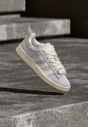 Baskets en daim violet clair avec des accents crème et des lacets blancs épais, flottant au-dessus de marches en béton dans un cadre extérieur.