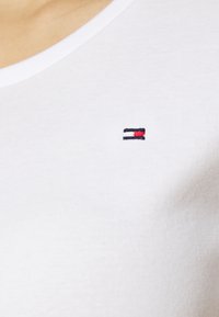 Biała bawełniana koszulka z dekoltem w serek, z małym haftowanym logo w kolorach niebieskim, białym i czerwonym umieszczonym po lewej stronie klatki piersiowej.