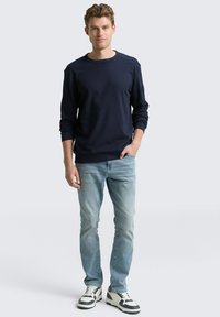 Marineblauwe long-sleeve crewneck sweatshirt met geribbelde manchetten, gecombineerd met lichtblauwe jeans en zwart-witte sneakers, staand tegen een lichte achtergrond.
