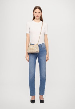 Beige Mini-Handtasche mit metallischem Netzdesign, goldenen Akzenten und einer Kettenriemen. Das Modell trägt ein enges weißes Oberteil und eine blaue Jeans mit schwarzen Ballerinas.