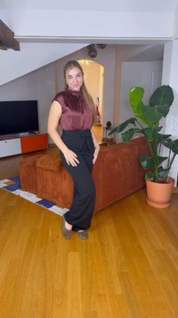 Burgundi blúz magas nyakkal, fekete, széles lábú nadrág bowval, fából készült padlón állva egy növény mellett és egy textúrált barna kanapéval.