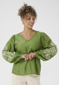 Blouse verte avec un décolleté en V, des manches bouffantes et des motifs tourbillonnants brodés en blanc sur les poignets. Fabriquée dans un tissu lisse et léger.