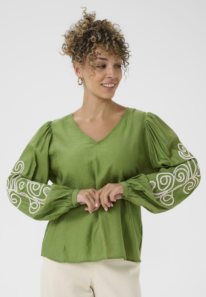 Blouse verte avec un décolleté en V, des manches bouffantes et des motifs tourbillonnants brodés en blanc sur les poignets. Fabriquée dans un tissu lisse et léger.