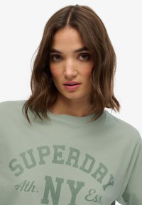 Sudadera de color verde menta, con cuello redondo y el texto "SUPERDRY" en verde oscuro. Tejido suave con un corte relajado y mangas cortas.