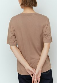 Smeđa pamučna majica s crew izrezom, kratkim rukavima i ravnim rubom. Tkanina djeluje mekano i glatko, s minimalnim detaljima šivanja.