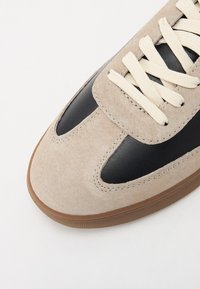 Sko med en beige semsket overdel med svarte skinnakenter, kremfargede snørebånd og såle i gummi, som viser et retro-design.