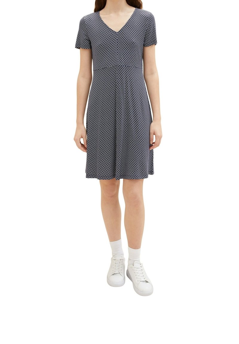 Femme portant une robe à manches courtes, longueur genoux, à motif noir et blanc, avec des chaussettes blanches et des baskets blanches, debout, bras détendus.