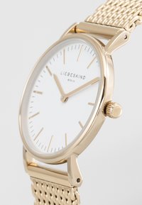 Gouden polshorloge met een witte wijzerplaat, minimalistische uurmarkers en een gaasband. Beschikt over een opvallende kroon en slanke designelementen.