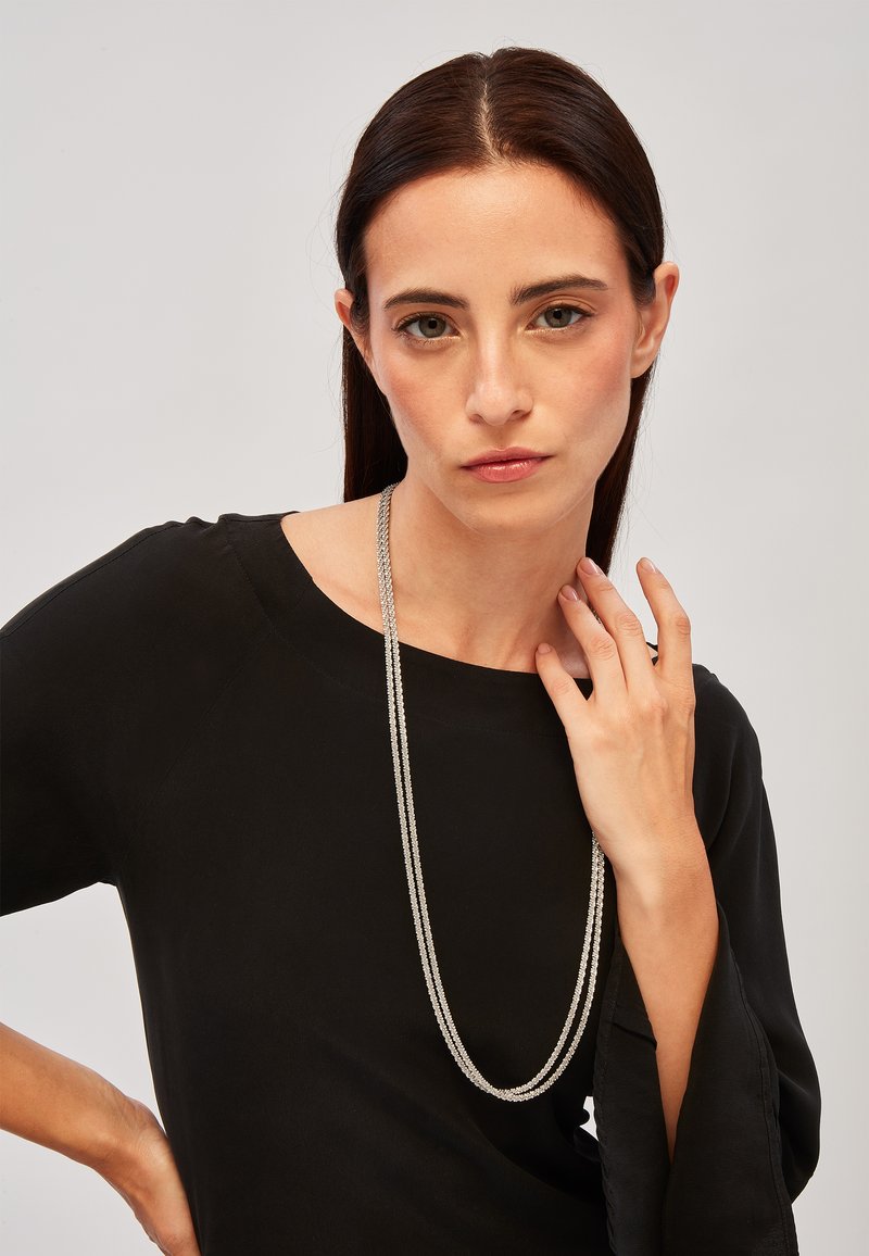 Sølvcirkelhalskæde med flere strenge, der hænger over en sort bluse. Modellen poserer med den ene hånd på halsen og viser halskædens længde og design.