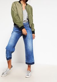 Oliwkowa bomberka z czarnymi akcentami, szara koszulka, jasnoniebieskie dżinsy w wersji cropped z postrzępionym dołem i srebrne sneakersy.