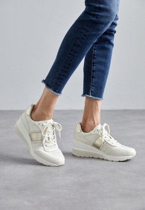 Osoba nosząca białe sneakersy na platformie z beżowymi akcentami i ciemnoniebieskie, skrócone dżinsy stojąca na szarej podłodze.