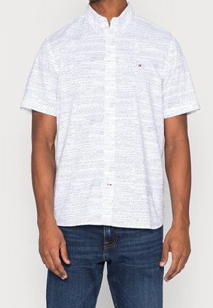 Mann trägt ein weißes kurzärmeliges Hemd mit kleinem schwarzem Punktmuster und dunkelblaue Jeans, steht vor einfarbigem Hintergrund.