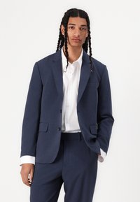 Traje azul marino con un blazer ajustado y pantalones, con una camisa blanca de botones debajo. Telas suaves con sutiles acentos de textura.