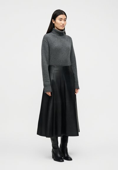Γυναίκα που φοράει γκρι πουλόβερ με ζιβάγκο, μαύρη midi φούστα από δέρμα με ψηλή μέση και μαύρα μπότες με τακούνι, στέκεται μπροστά σε απλό φόντο.