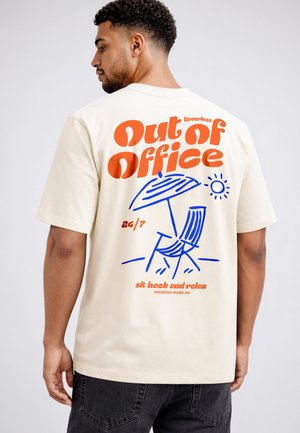 Man wearing cream T-shirt with orange text "Out of Office" and blue beach chair, umbrella, and sun illustration on back.