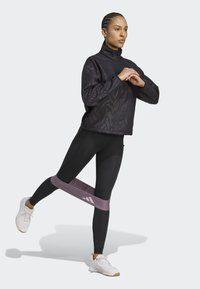 Pull noir à fermeture éclair avec un motif texturé, associé à un legging noir et une bande de résistance violette claire. Des baskets blanches complètent le look.