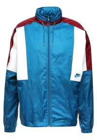 Veste Nike au design surdimensionné en bleu, blanc et maroon. Fabriquée en tissu brillant, avec fermeture éclair et deux poches latérales.