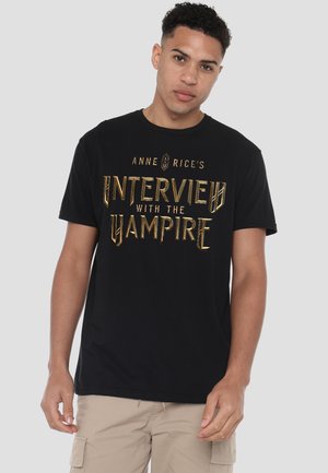 Junger Mann trägt ein schwarzes T-Shirt mit goldenem Text "Anne Rice's Interview with the Vampire" und beige Cargoshorts, steht vor einem einfarbigen Hintergrund.