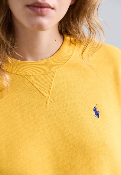 Sudadera amarilla con cuello redondo acanalado, que presenta un pequeño logo bordado azul en el pecho. Tejido suave con una textura lisa.