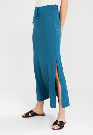 Frau trägt einen türkisfarbenen, knöchellangen Rock mit Seitenschlitz und Kordelzug in der Taille, kombiniert mit schwarzen flachen Sandalen mit gekreuzten Riemen.