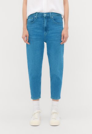 Person trägt blaue hoch taillierte kurze Jeans, weiße Socken, weiße Sneakers und ein reingestecktes weißes Oberteil und steht vor einem einfarbigen Hintergrund.