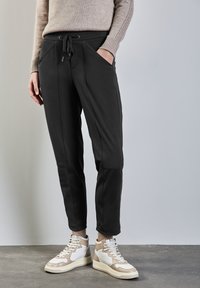 Pantalones jogger negros con cintura elástica, bolsillos laterales y corte ajustado. Combinados con zapatillas altas en beige y blanco. Textura suave.