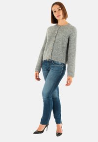 Cardigan de lana gris claro, con cuello redondo, mangas largas y botones en la parte delantera. Combinado con jeans ceñidos azules y tacones negros.