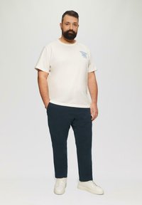 Beige katoenen T-shirt met blauwe tekst, marineblauwe broek en witte sneakers. Model staat met handen in de zakken voor een effen achtergrond.