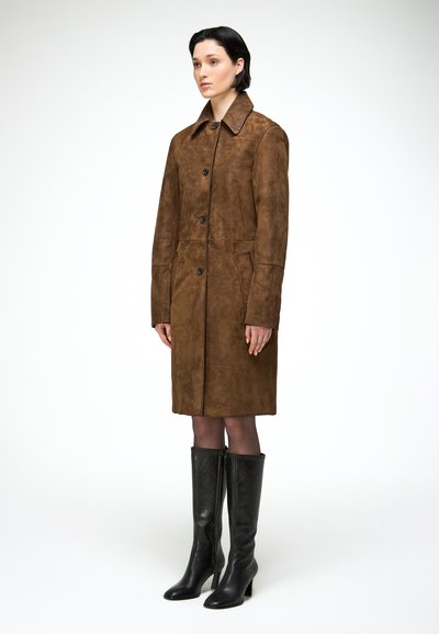 Femme aux cheveux courts et foncés portant un manteau en daim marron longueur genou et des bottes à talons noires montant jusqu'au genou, posant devant un fond blanc.