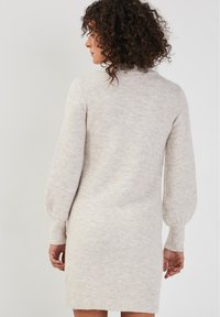 Robe en tricot beige doux avec un col montant, de longues manches bouffantes et des poignets côtelés. Présente une coupe décontractée et tombe au-dessus des genoux.