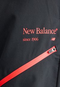 Sort stof med rød "New Balance siden 1906" tekst og logo over et diagonalt rødt lynlåsdetalje.