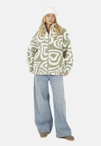 Fleece zip-up jack in lichtgroen met witte abstracte patronen; gecombineerd met losse blauwe denim jeans en een witte beanie.