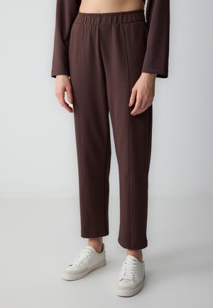 Person trägt braune gerade geschnittene Hose mit elastischem Bund und weiße Turnschuhe, steht vor einem schlichten hellen Hintergrund.