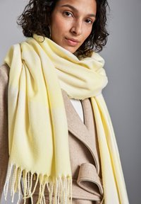 Sciarpa gialla morbida realizzata in un tessuto texturizzato, con estremità frangianti, disposta elegantemente attorno al collo, abbinata a un cappotto beige.